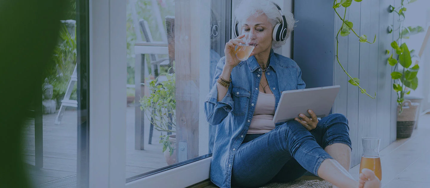 A woman with headphones and a tablet sitting next to a window with a refreshing drink.